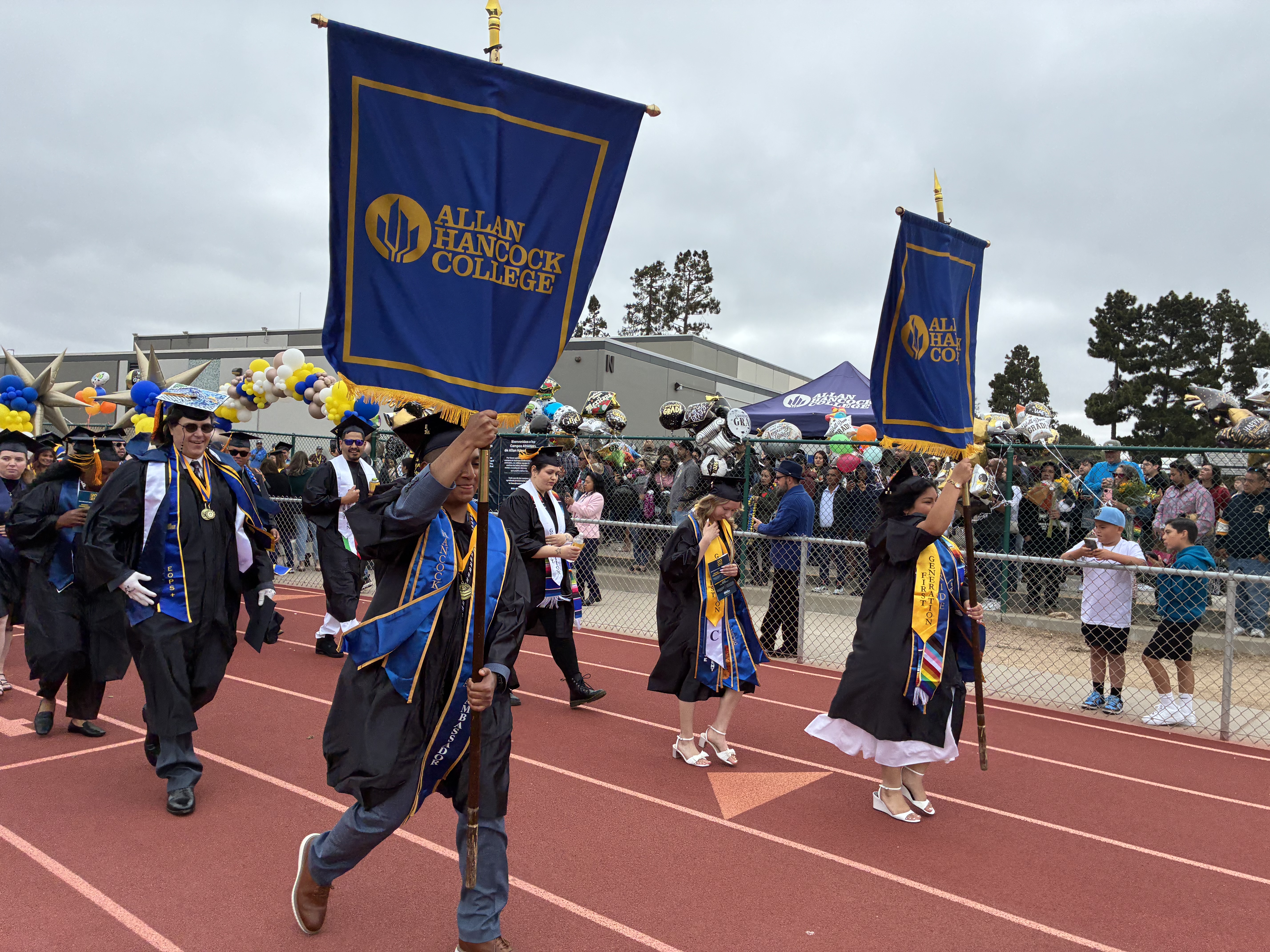 Graduates carry Allan Hancock College banners while leading the procession during the college’s 2026 commencement ceremony. Wearing caps, gowns, medals, and honor stoles, the graduates walk along a track as family members and supporters cheer from behind a fence decorated with balloons and celebration signs
